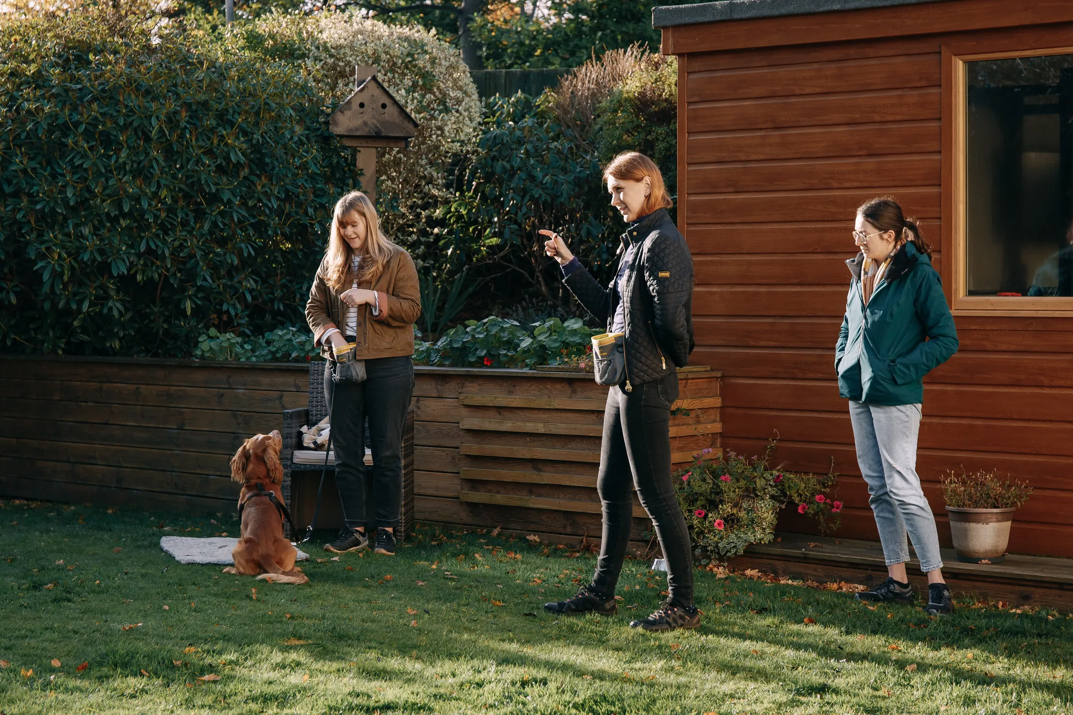 Katya coaching two owners during a puppy class in a sunny Colinton garden, a spaniel puppy sitting on a mat